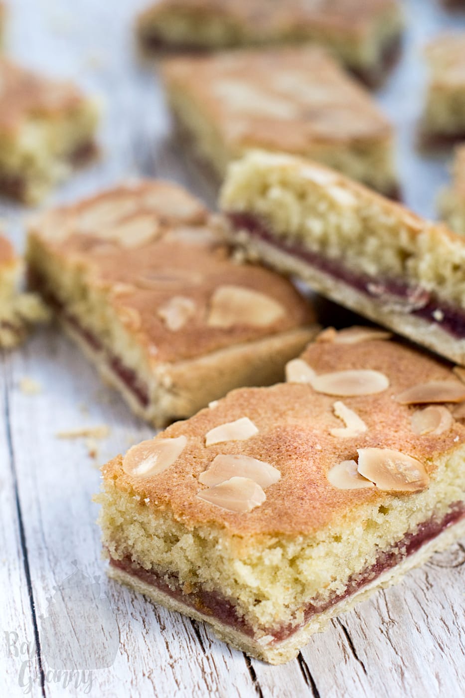 Close-up shot of a sliced homemade Almond tray bake, showing distinct layers of shortcrust pastry, dark raspberry jam, and a thick almond frangipane sponge topped with golden flaked almonds.