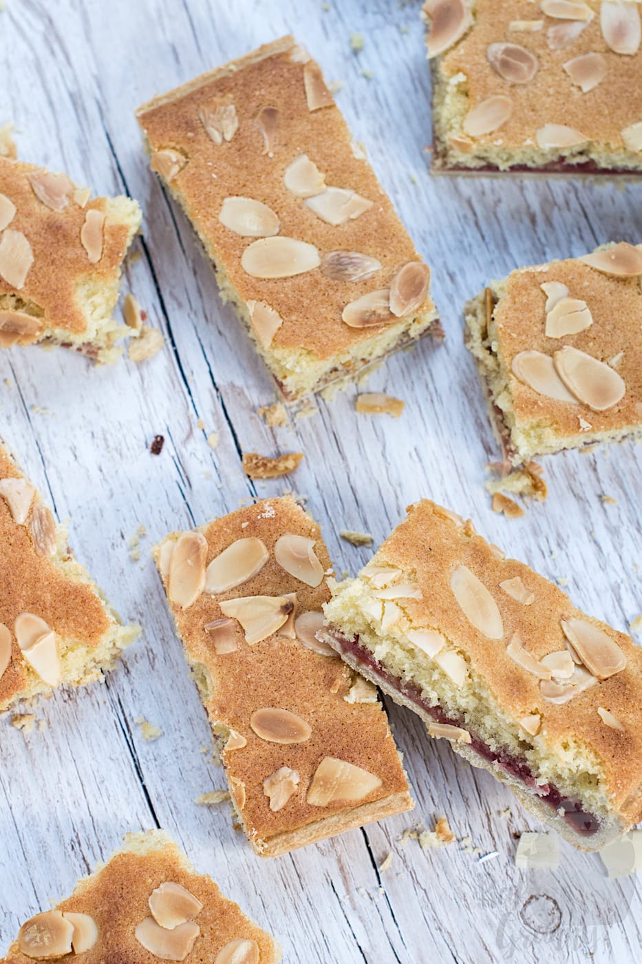 Flat lay overhead view of several sliced Bakewell tray bake pieces scattered on a light-coloured wooden background with toasted almond flakes around them.