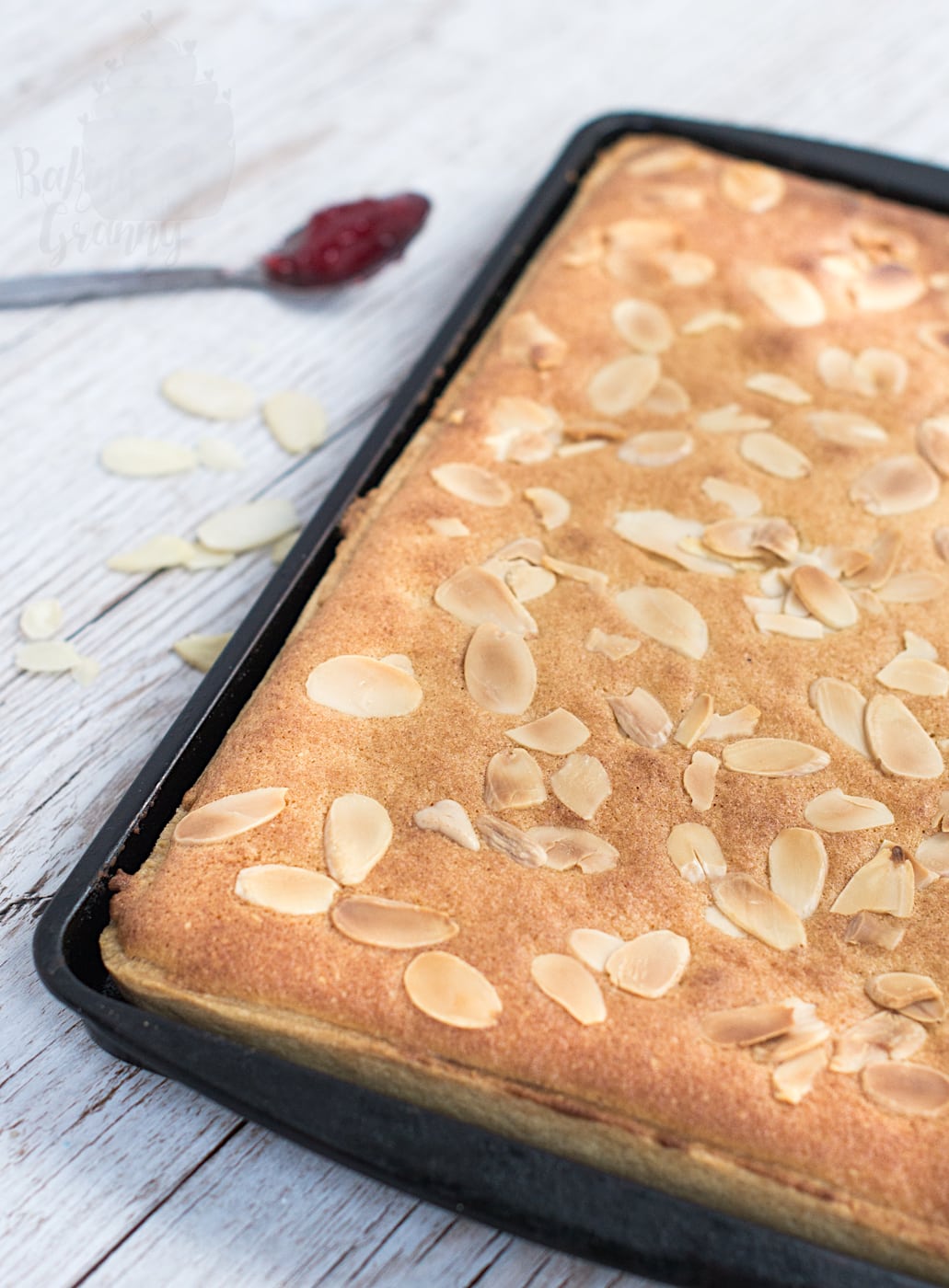 A freshly baked almond slice tray bake still whole inside a rectangular metal baking tin, showing a golden frangipane surface covered in toasted flaked almonds with a spoonful of jam in the blurry background.