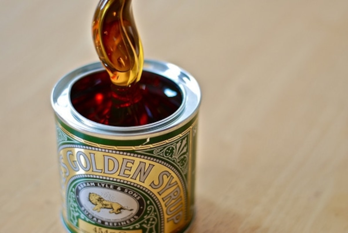 A classic green metal tin of golden syrup with a silver spoon lifting a thick, glossy strand of sticky amber syrup upwards.
