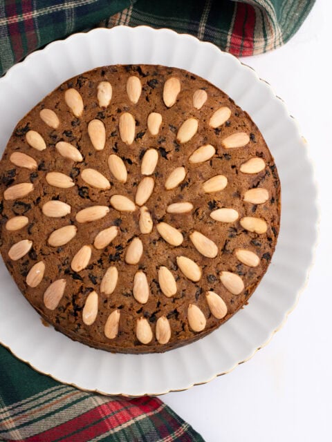 Traditional Scottish Dundee Cake, with almond topping, packed with rich dried fruit. On a white plate, atop a tartan fabric.