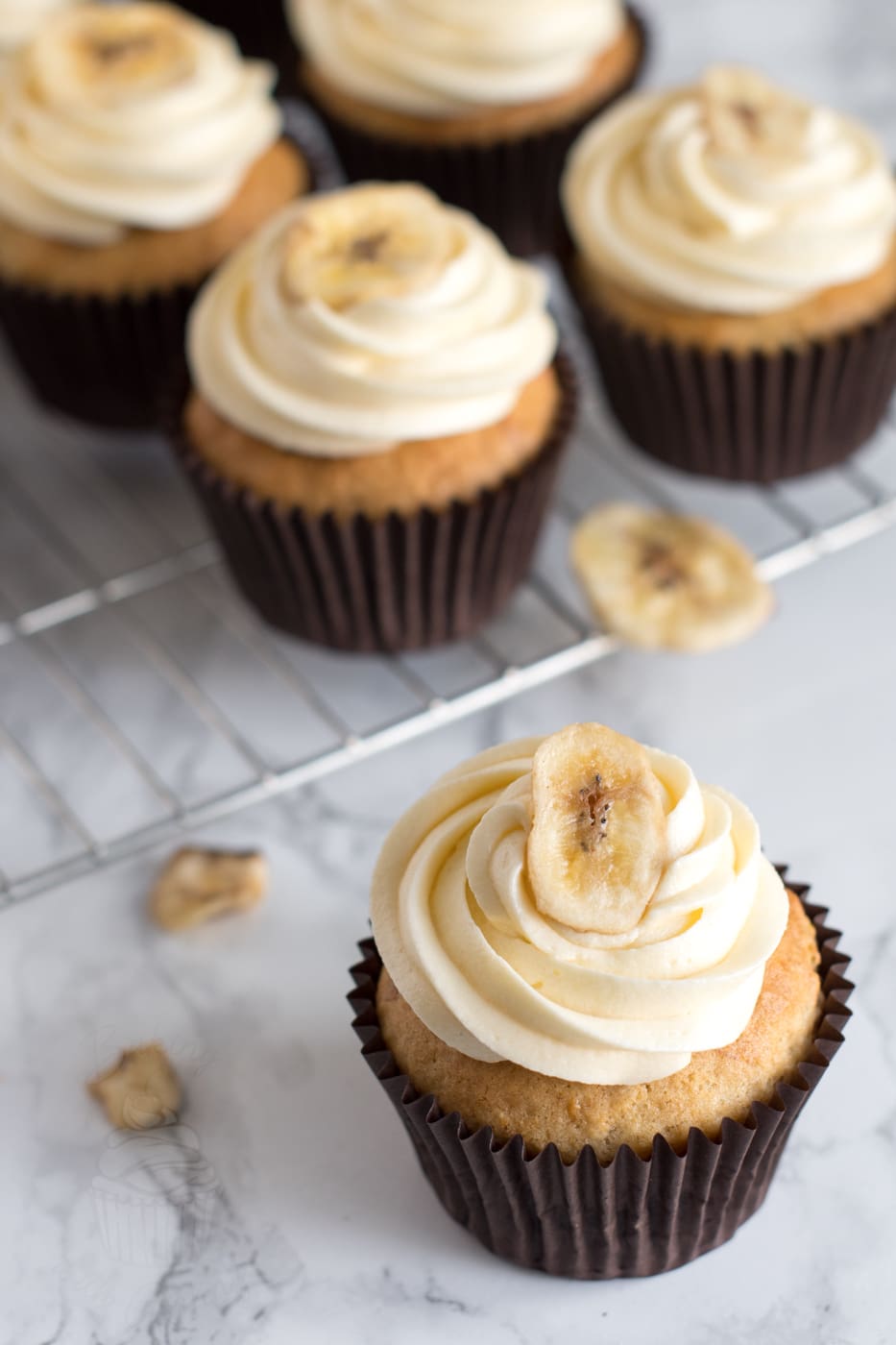 Banana Cupcake topped with vanilla buttercream and a dried banana chip, with more cupcakes cooling in the background.