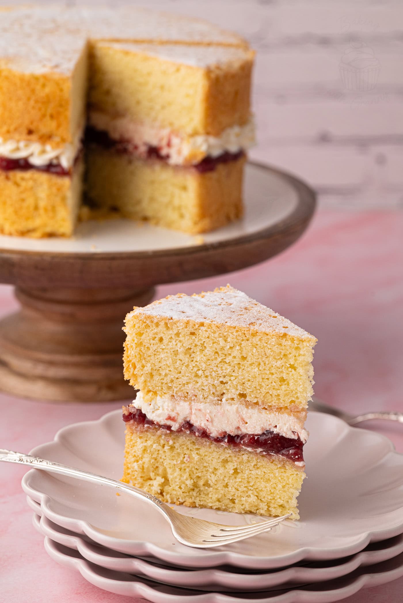 Slice of Victoria Sponge showing sponge, strawberry jam and buttercream, with the remaining cake behind.