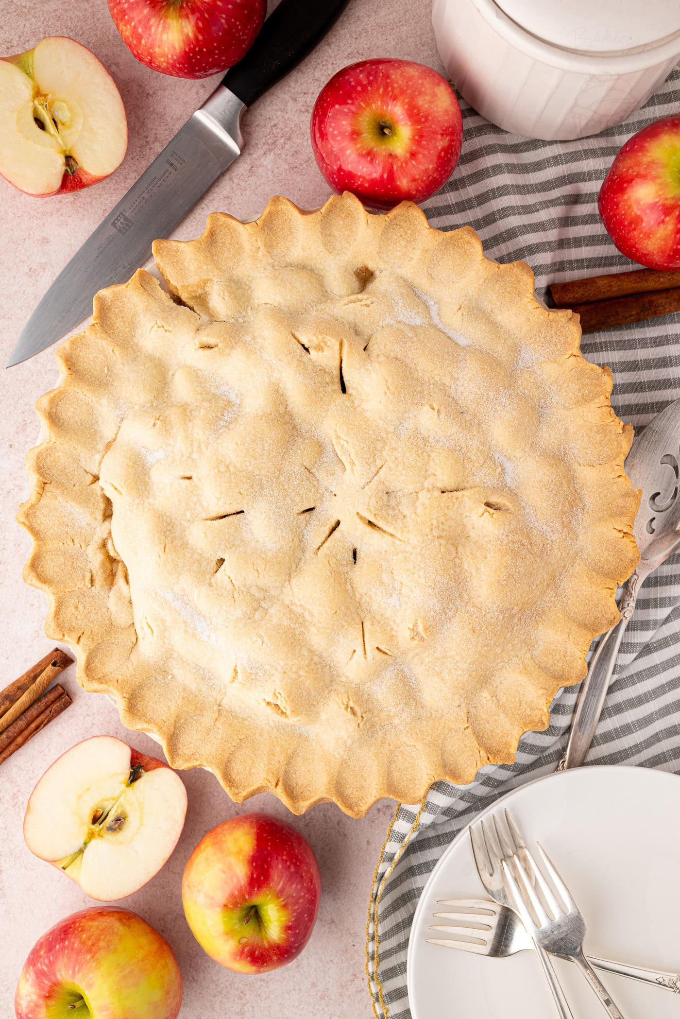 Whole baked apple pie with apples, cinnamon sticks and cutlery around it.