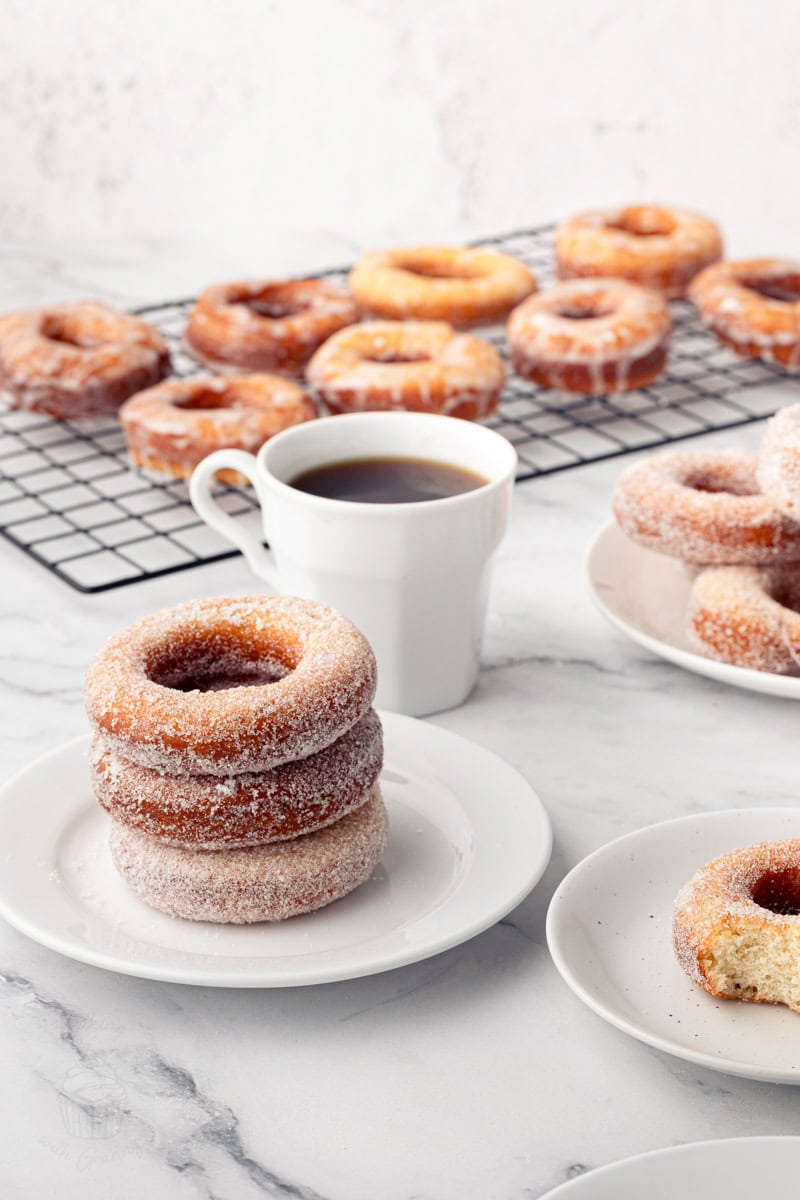 A table setting featuring a stack of sugared donuts, a white coffee cup, and a wire cooling rack covered in glazed ring donuts in the background.