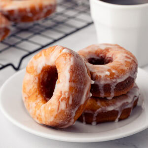 A plate of fresh homemade ring donuts covered in a dripping white sugar glaze, served next to a mug of coffee.