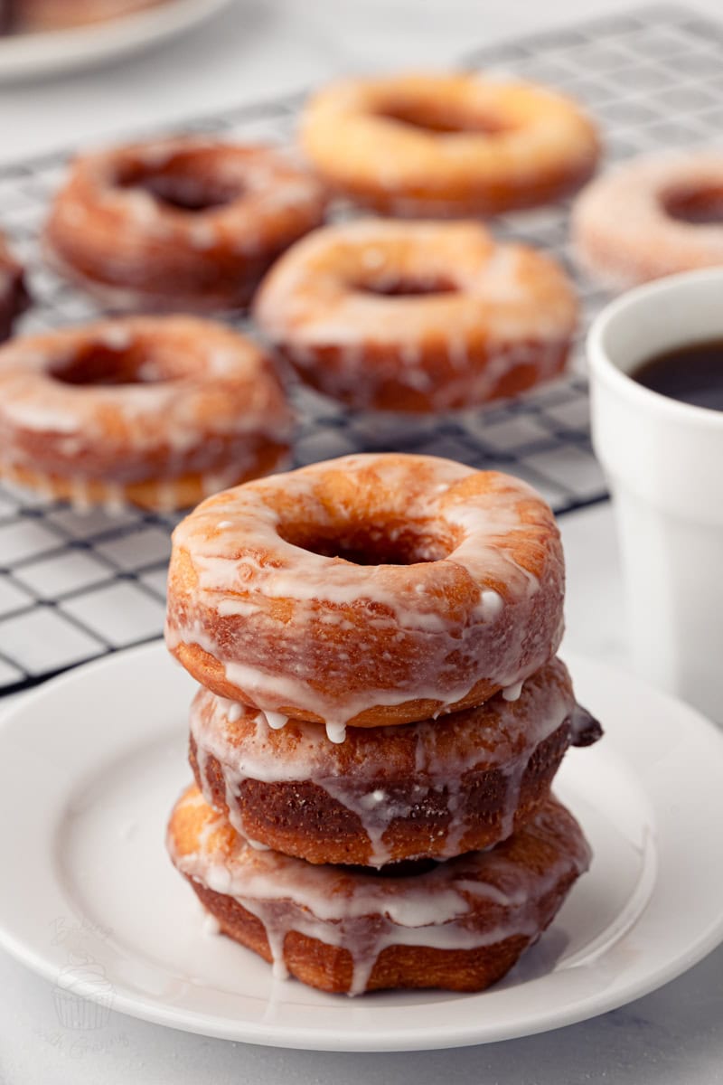 A stack of three glossy, glazed homemade ring donuts on a white plate, with more donuts cooling on a wire rack behind them.