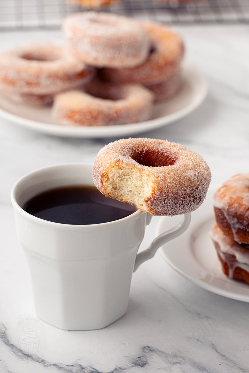 A fun shot of a sugar-coated ring donut with a bite missing, balanced on the rim of a white mug filled with black coffee.
