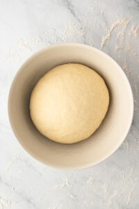 A smooth ball of homemade doughnut dough proofing inside a large ceramic bowl on a floured marble surface.