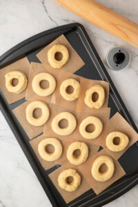 Risen, unbaked ring doughnuts on parchment paper squares spread across a baking tray, ready to be fried.