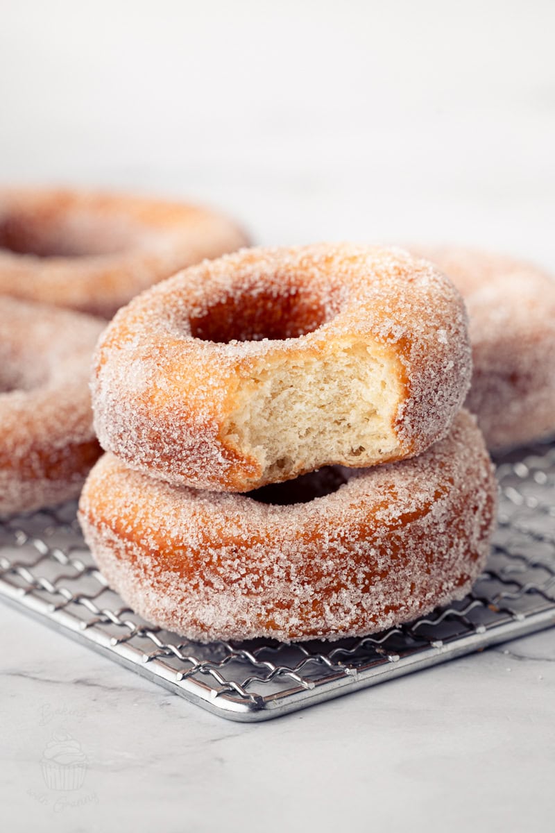Two sugar-crusted ring doughnuts stacked on a wire cooling rack, with the top one showing a large bite taken out.