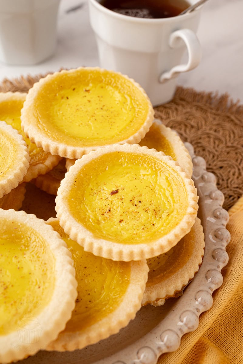 Plate stacked with traditional British custard tarts, served with a cup of tea in the background.