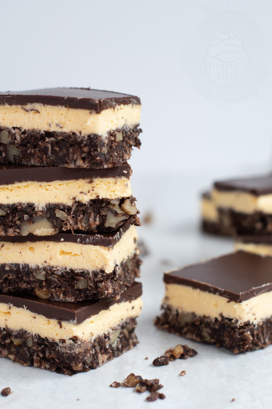 A tall stack of four homemade Nanaimo bars showing the distinct three layers of chocolate, custard cream, and coconut-biscuit base, with crumbs on a white surface.