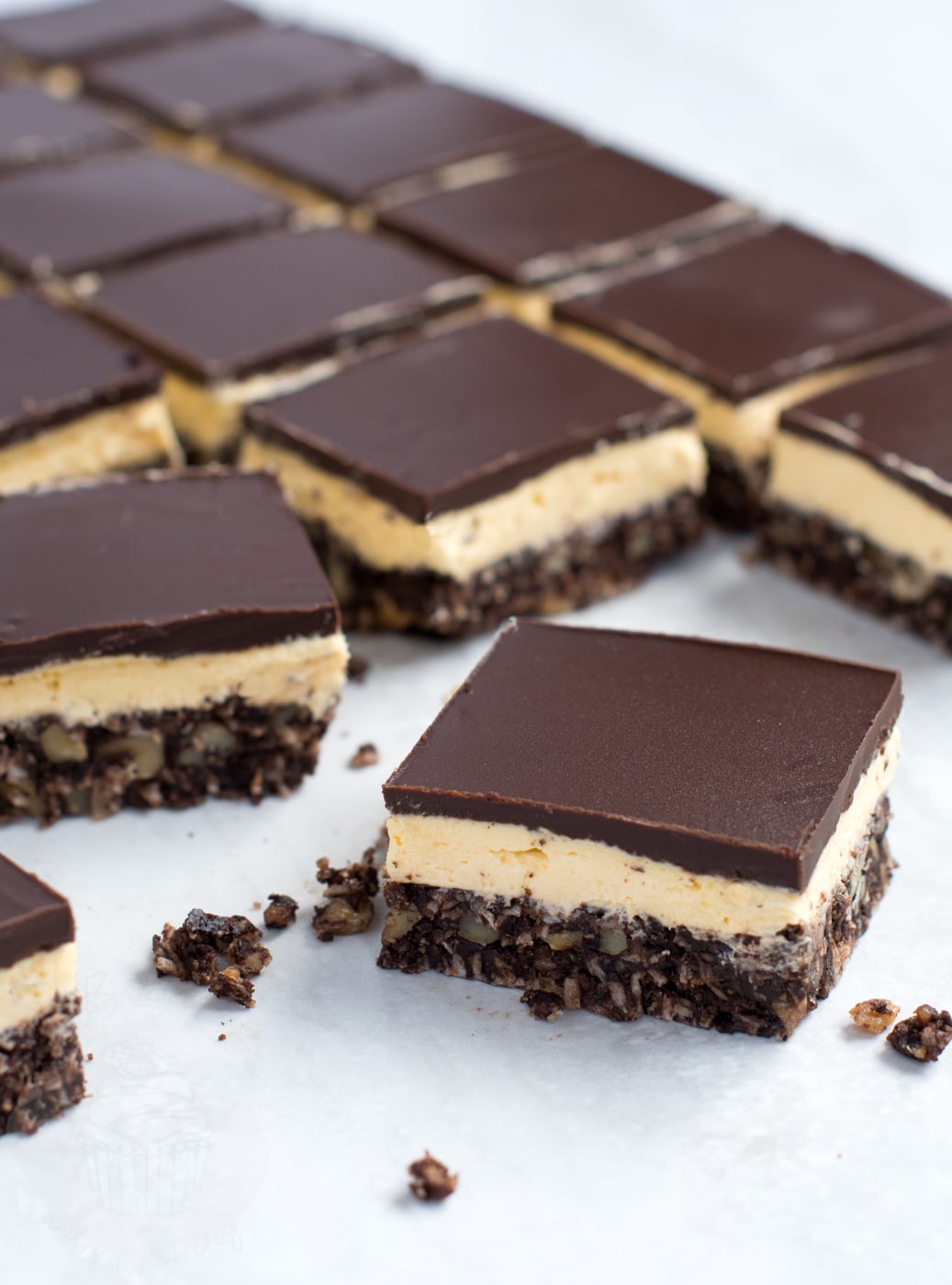 A single square Nanaimo bar slice in the foreground with a grid of neatly sliced chocolate custard bars lined up behind it.