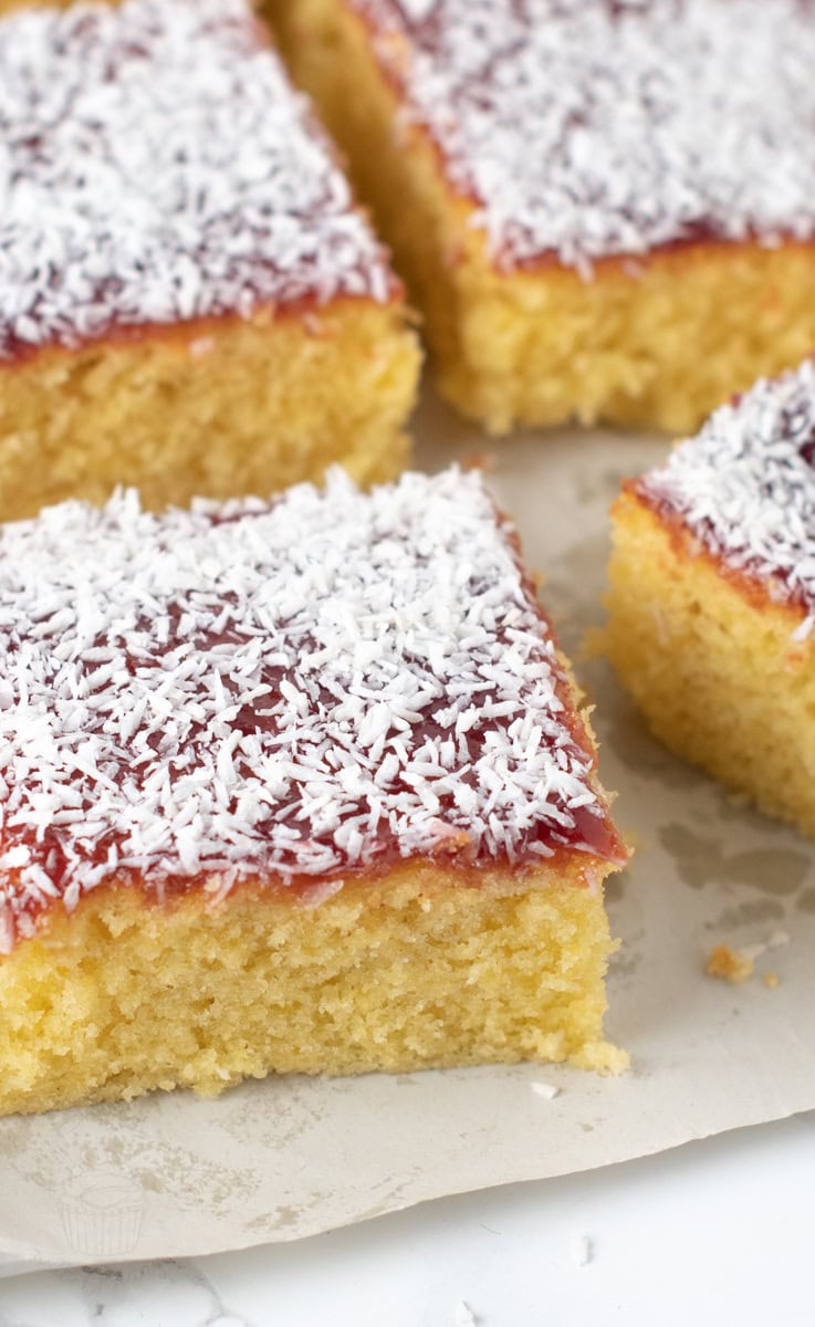 Slices of jam and coconut school cake on baking parchment showing the yellow sponge beneath the jam and coconut topping.