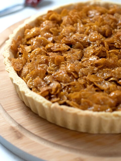 Close up of a golden cornflake tart, on a round wooden board, with a spoonful of jam in the background.