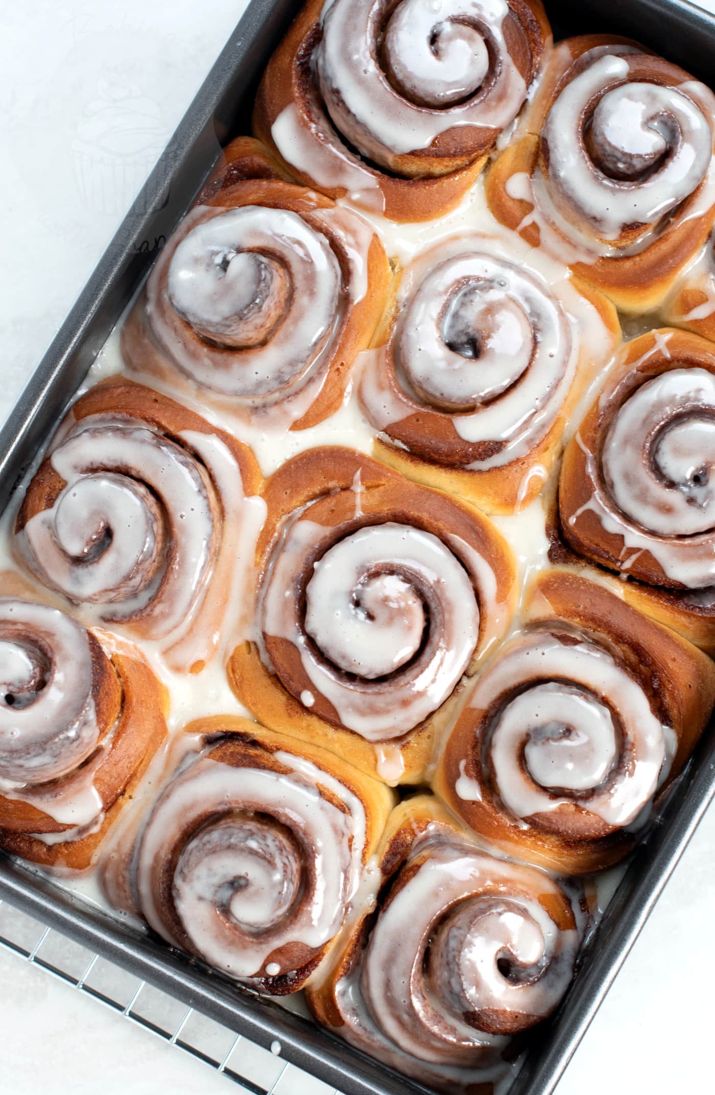 Tin of freshly baked and iced Cinnamon Buns in a tray, on top of a wire cooling rack.