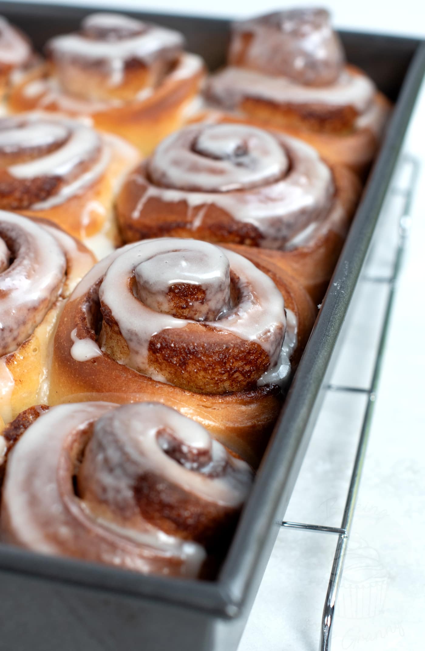 Homemade Cinnamon Rolls cooling in a metal tin, on top of a wire cooling rack, with fresh white icing on top of the buns.