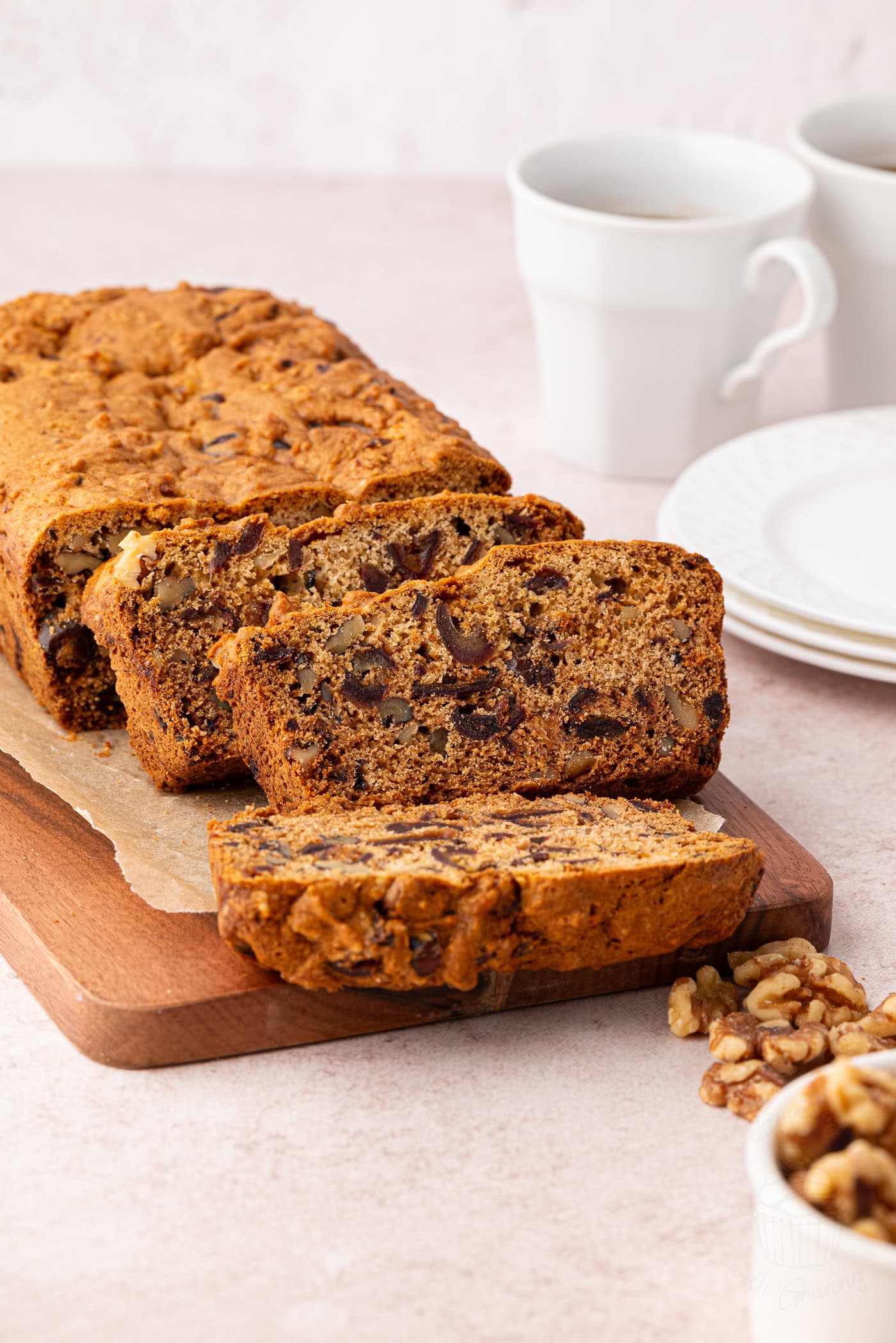 Sliced walnut and date loaf on a wooden cutting board with two coffee cups and saucers in the background.