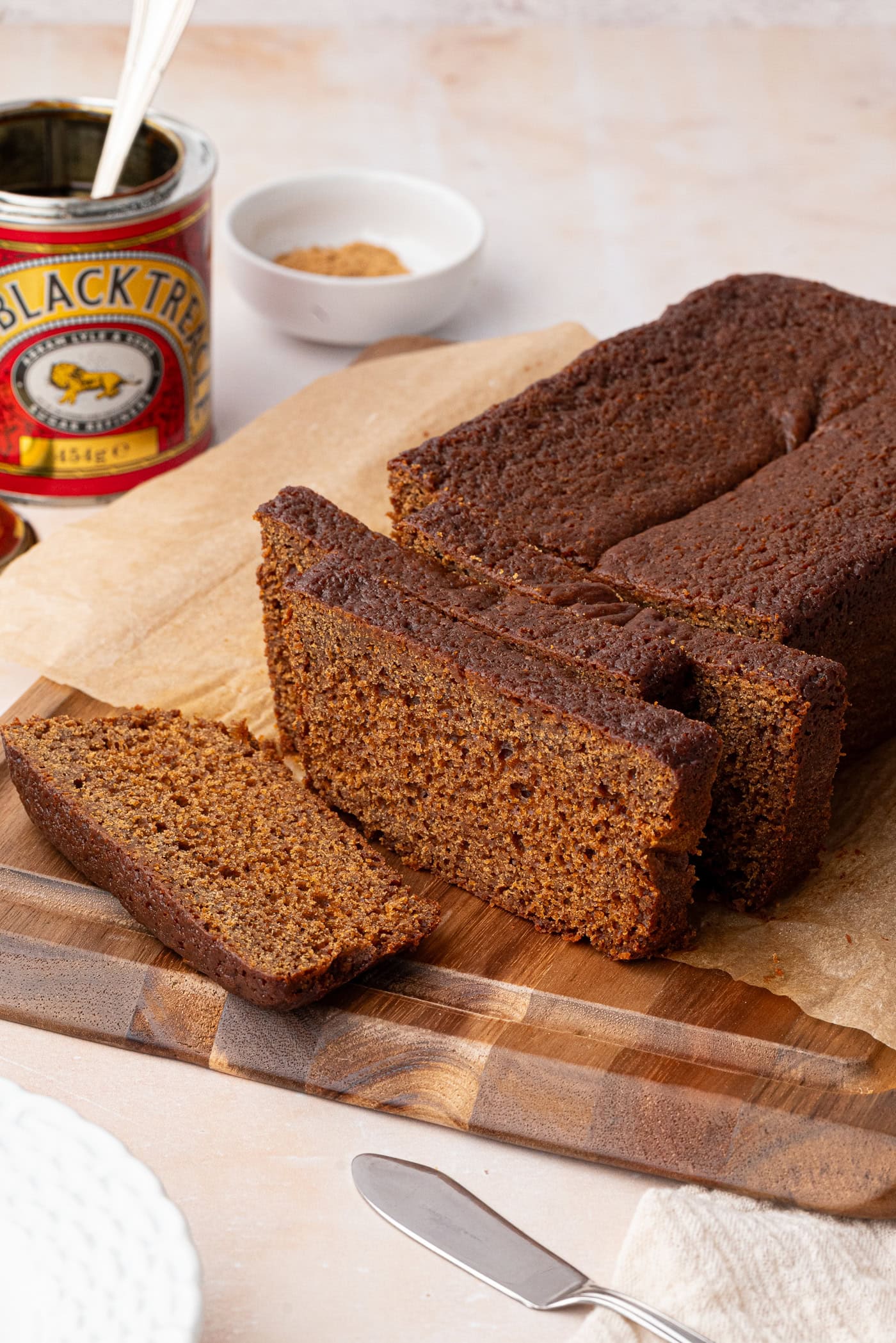 Sliced sticky gingerbread loaf on a wooden board beside treacle and ground ginger.