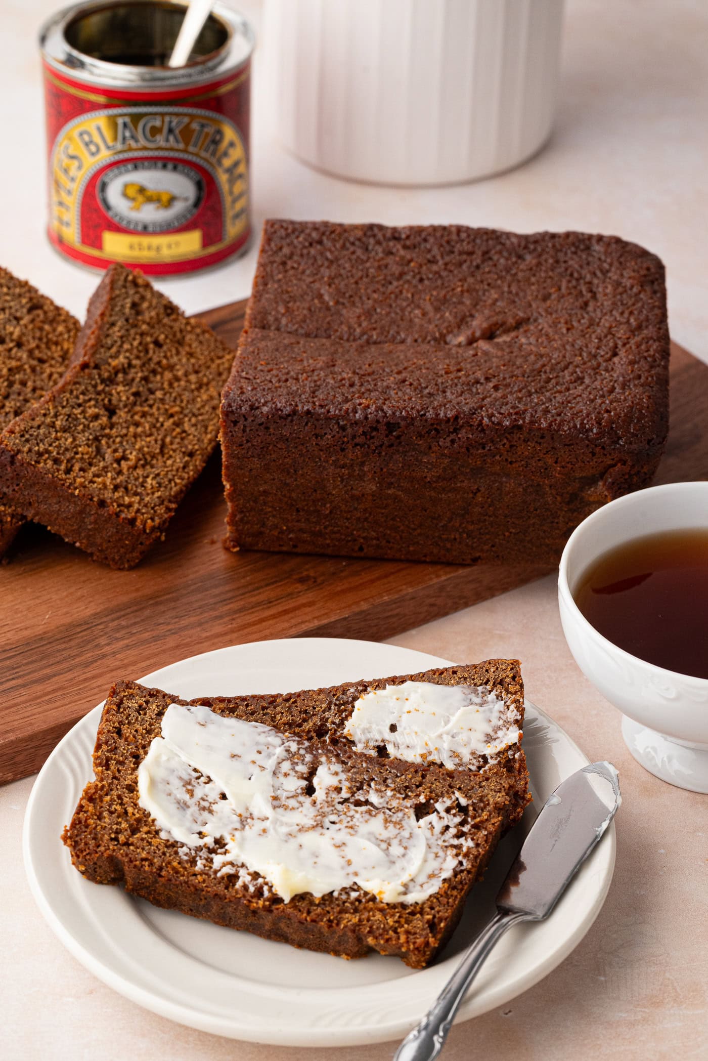 Slice of gingerbread loaf spread with butter beside a cup of tea.
