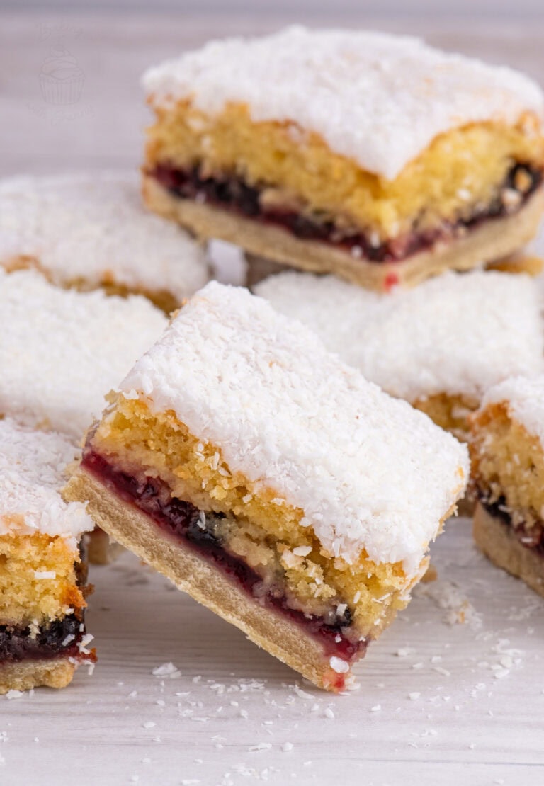 Scottish Snow Cake slices stacked on a white surface, showing the jam and sponge layers beneath a dusting of coconut.