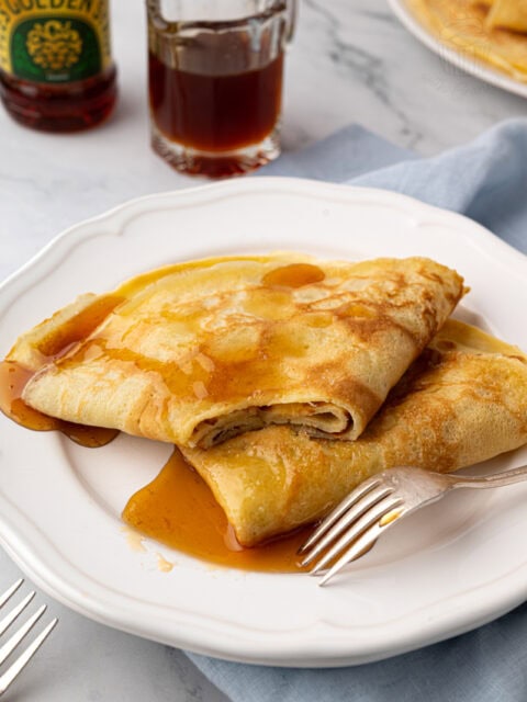 Two folded pancakes drenched in rich golden syrup on a white plate, with a silver fork resting on the side.