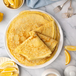 An overhead view (flat lay) showing a stack of thin, large round British pancakes on a white plate, surrounded by bowls of lemon slices, a sugar pot, and two silver forks.