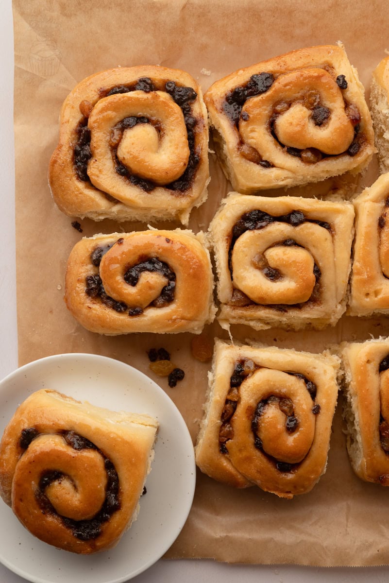 Overhead view of homemade Chelsea Buns on baking paper with one bun served on a white plate.