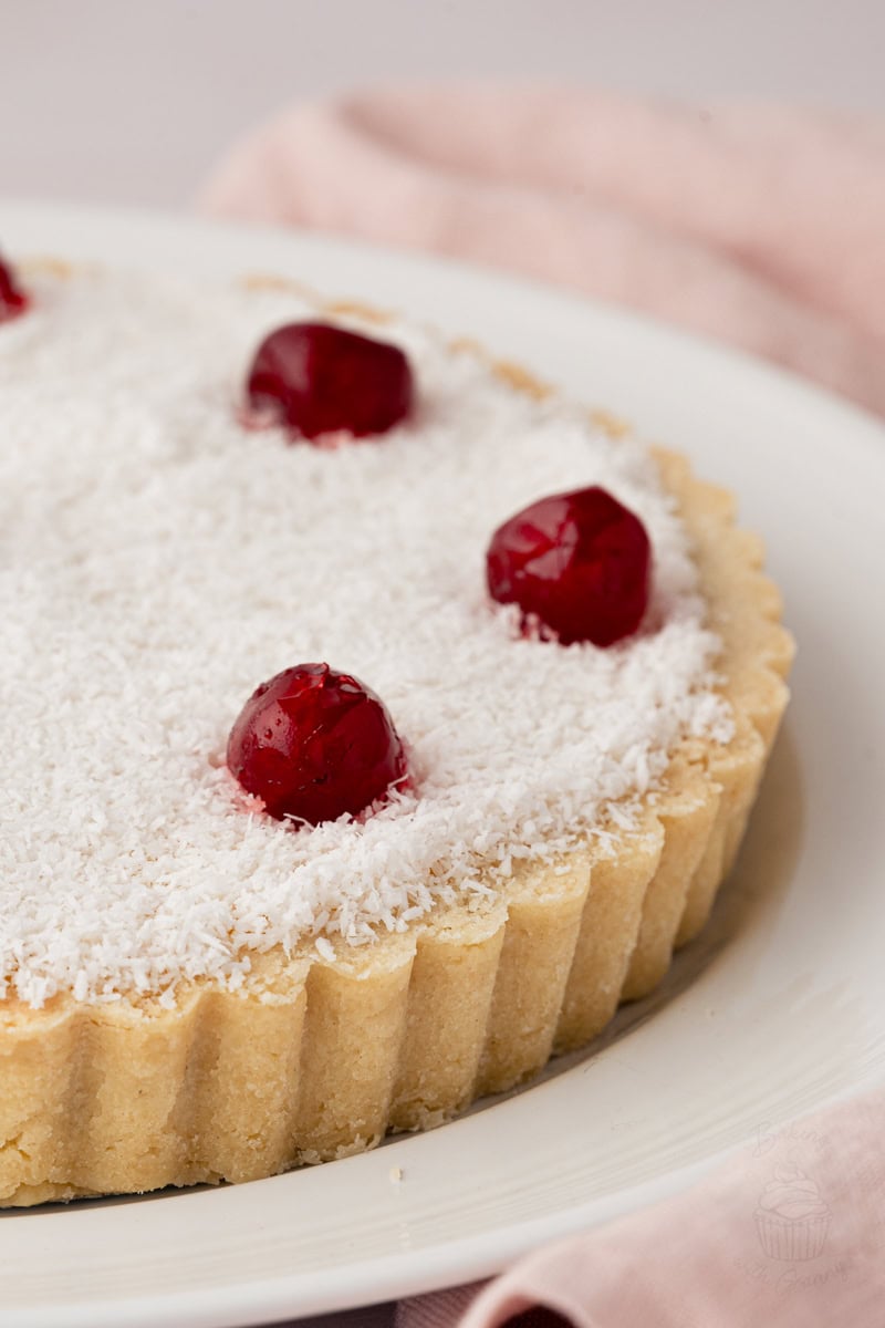 Close-up of homemade Manchester Tart with a buttery pastry case, creamy custard filling, coconut topping and glace cherries.