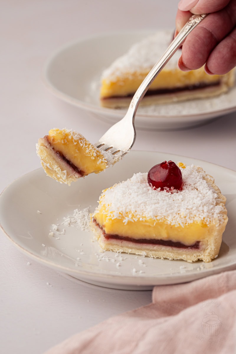 Fork lifting a bite of homemade Manchester Tart with creamy custard, raspberry jam and coconut topping from a slice on a dessert plate.