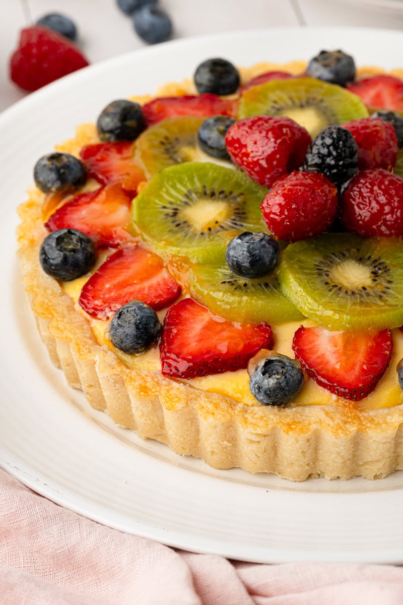 Close-up angled shot focusing on the golden, fluted pastry crust of a homemade fruit tart, topped with glossy, apricot-glazed kiwi, strawberries, and blueberries.