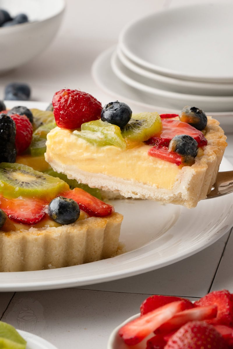A silver pie server lifting a slice of fresh fruit tart from the main pastry shell, showcasing the thick layer of vanilla custard cream and the vibrant fruit topping.