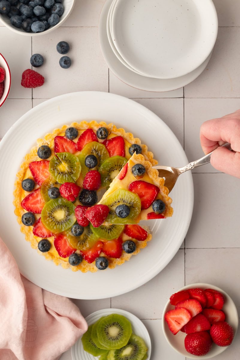 Flat lay overhead view of a baker's hand using a silver server to lift a slice from a whole glazed fruit tart, surrounded by decorative bowls of berries and kiwi slices.