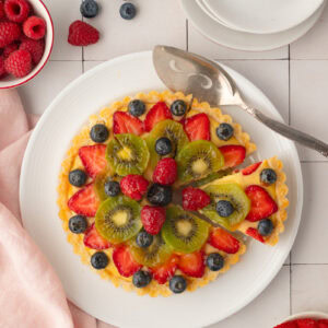 Symmetrical top-down view of a colorful summer fruit tart with one slice gently separated from the rest, highlighting the neat, fluted edges of the shortcrust pastry.