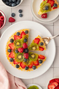 Top-down view of a classic French fruit tart on a white plate, with a silver cake server lifting out a single slice to show the colorful geometric pattern of kiwi, strawberries, and blueberries.