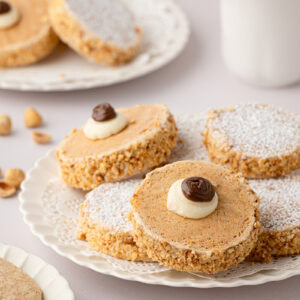 Close-up shot of a plate of traditional Scottish Japs biscuits, highlighting the ground hazelnut coating on the buttercream sides and a piped buttercream decoration with a chocolate dollop on top.