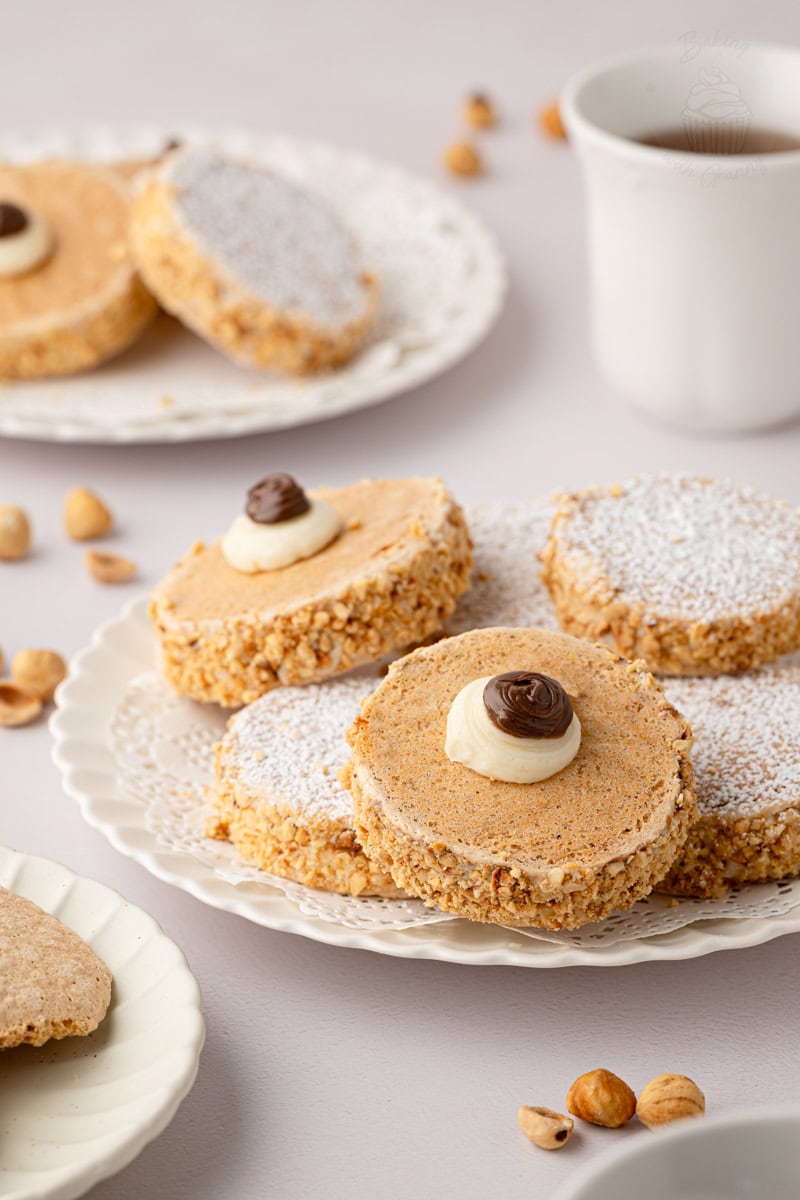 Close-up shot of a plate of traditional Scottish Japs biscuits, highlighting the ground hazelnut coating on the buttercream sides and a piped buttercream decoration with a chocolate dollop on top.