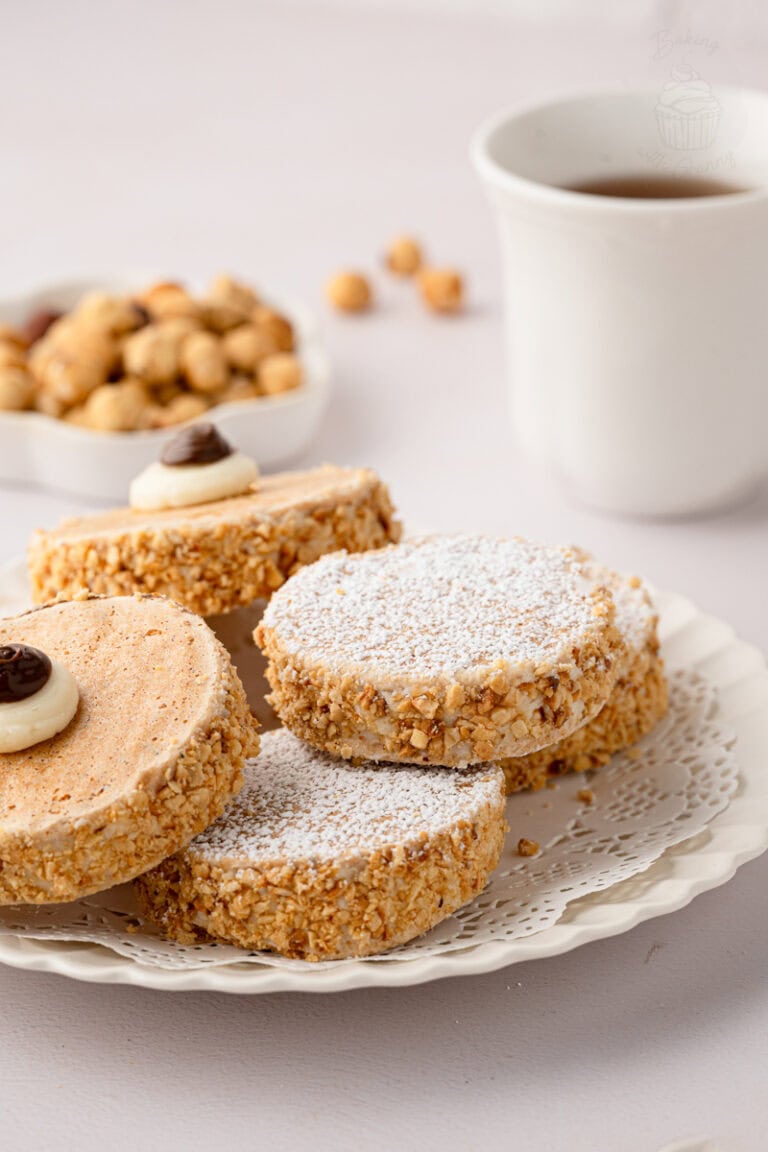 Eye-level close-up of a stacked pile of icing-sugar-dusted Japonaise cakes on a white plate with a vintage lace doily.