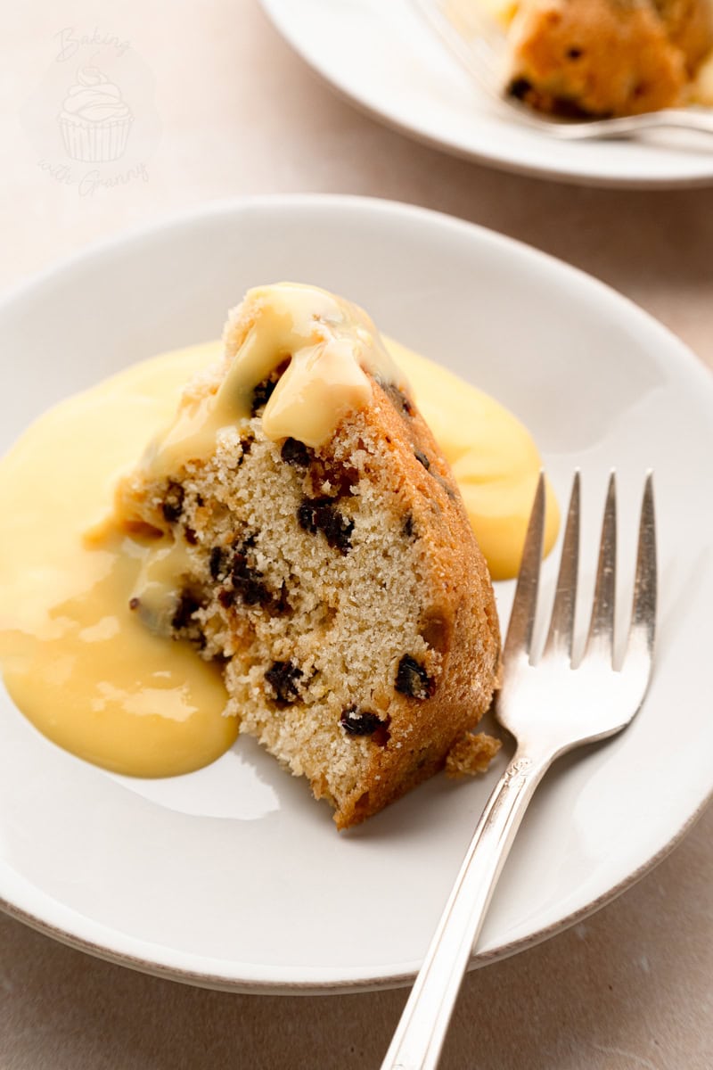 Top-down view of a slice of currant-spotted steamed pudding topped with custard, served with a vintage silver fork.