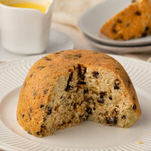 A steamed Spotted Dick pudding on a plate with a large slice cut out, revealing the soft sponge interior and dried fruit.