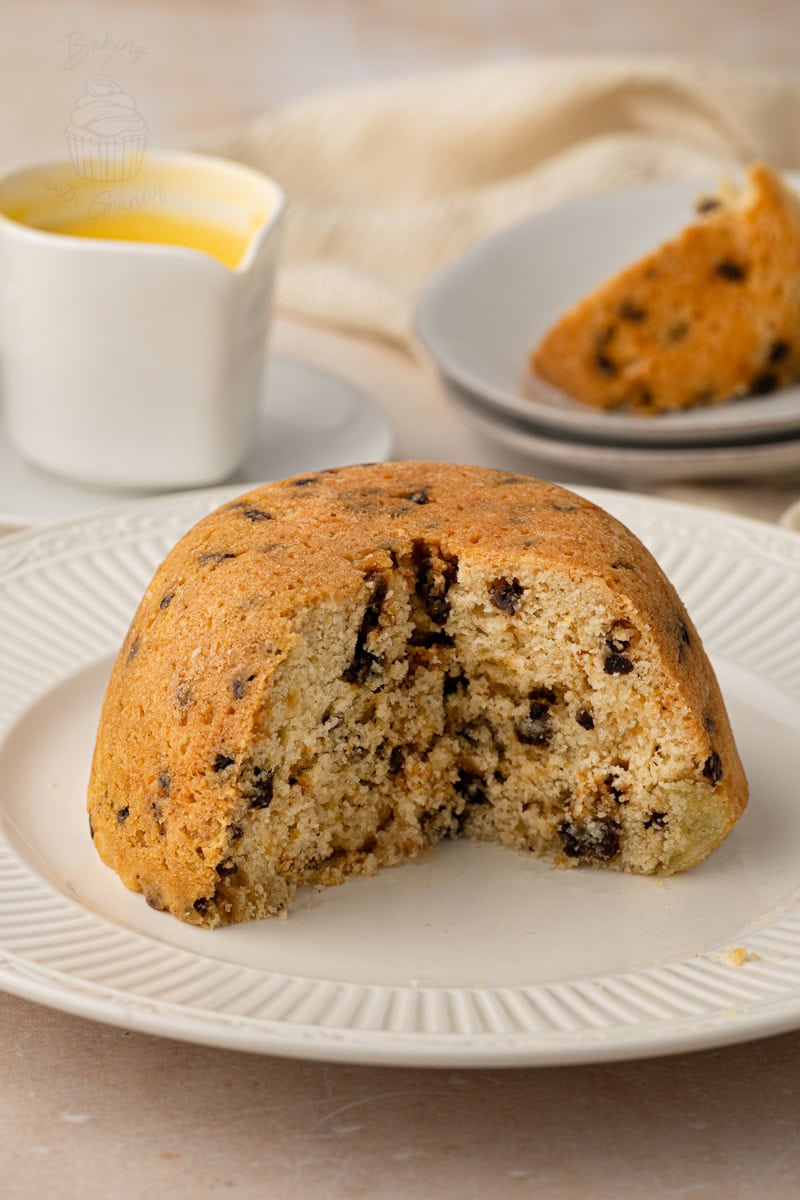 A steamed Spotted Dick pudding on a plate with a large slice cut out, revealing the soft sponge interior and dried fruit.