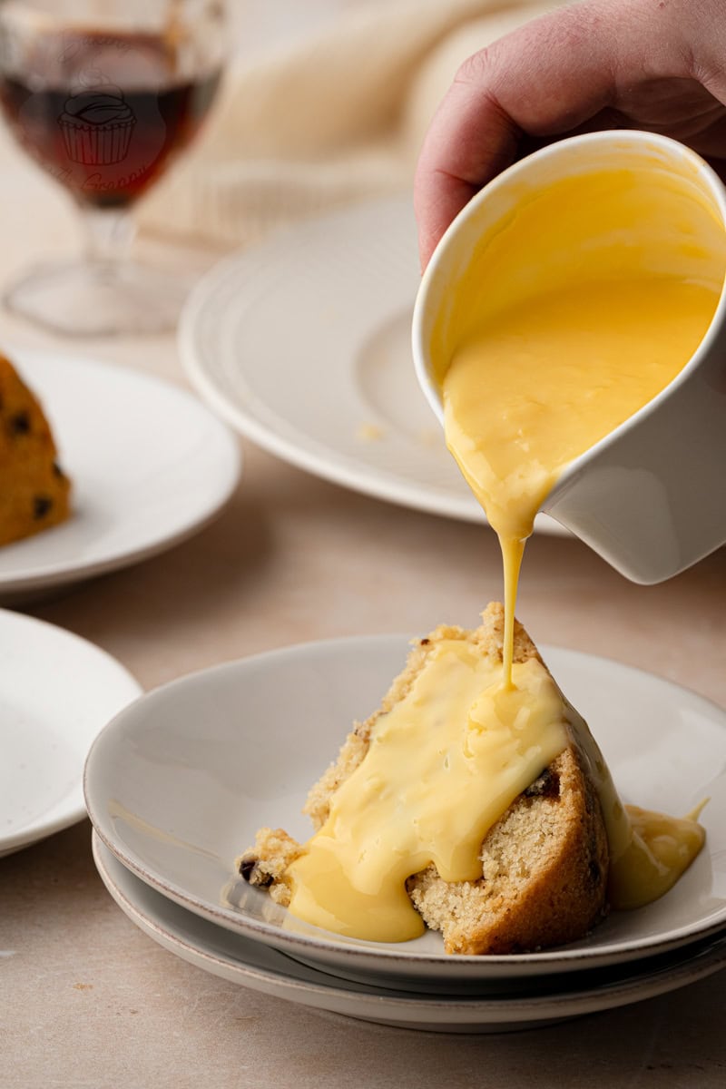 Warm yellow custard being poured from a white jug over a slice of traditional Spotted Dick pudding.