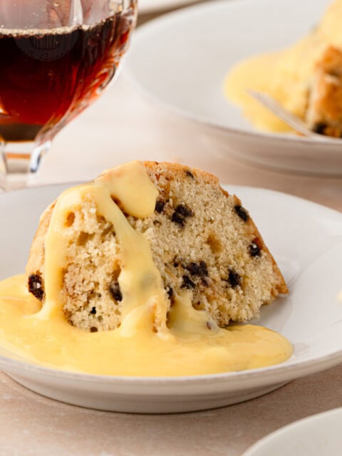 A slice of classic British Spotted Dick pudding sitting in a pool of smooth yellow custard.