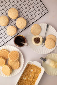 Overhead view of the baking process for Scottish Japonaise cakes, featuring a jar of chocolate spread, piped buttercream on a shell, a cooling rack, and a dish of ground hazelnuts.