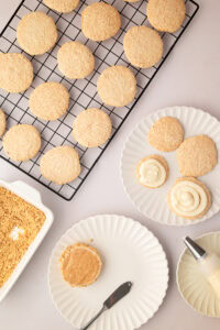 Step-by-step assembly of hazelnut meringue biscuits, showing buttercream piped in a spiral onto the flat side of the shells next to a dish of chopped hazelnuts.
