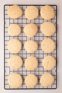Row of freshly baked, golden-brown hazelnut meringue biscuit shells cooling on a black wire baking rack.