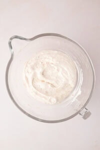 Whipped egg white meringue batter folded with ground hazelnuts in a large glass stand mixer bowl, showing a thick and glossy texture.