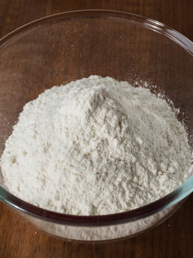 A clear glass mixing bowl filled with a neat mound of white self-raising flour sitting on a wooden kitchen surface.