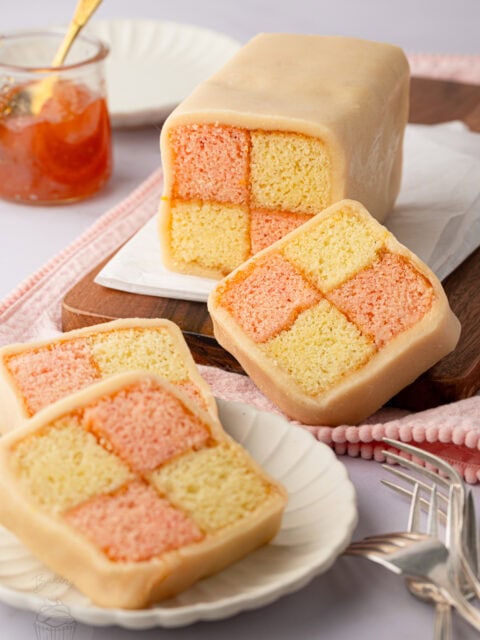 Whole Battenberg cake and sliced pieces showing pink and yellow checkerboard sponge wrapped in marzipan, with apricot jam beside it.